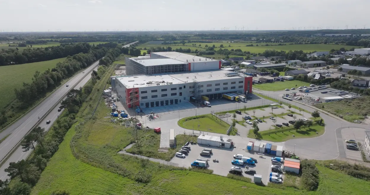 Eine Luftaufnahme eines modernen Lagergebäudes mit grauer Fassade und roten Akzenten. Vor dem Gebäude sind Lkw und Lieferwagen im Parkbereich zu sehen. Umgeben von Wiesen und einer Straße, während im Hintergrund Windräder sichtbar sind.