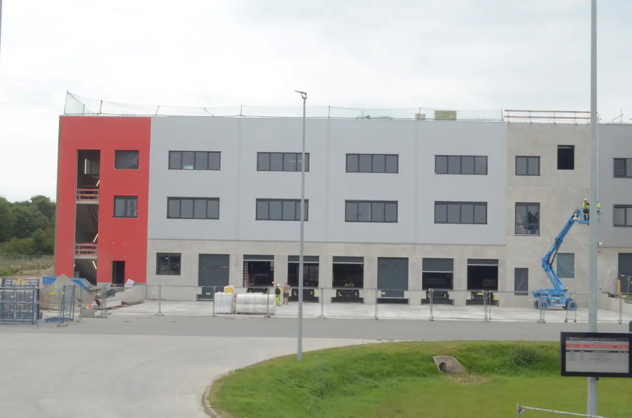 Ein im Bau befindliches Gebäude mit einer roten und grauen Fassade. Auf der Baustelle sind Bauarbeiter und ein Arbeitslift zu sehen. Im Vordergrund befinden sich Baustellenmaterialien und eine Umzäunung, während eine Straße und Rasenflächen den Hintergrund bilden.