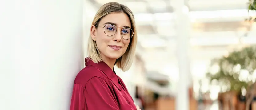Junge Frau mit Brille lehnt entspannt an einer Wand im hellen Büro.
