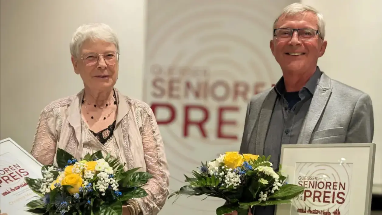 Preisträger und Preisträgerin des Queisser Seniorenpreises 2024 mit Blumen und einer Urkunde vor einem Roll-Up