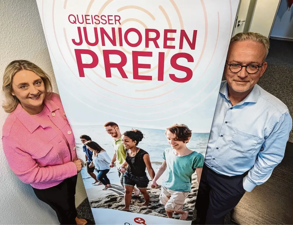 Zwei Personen stehen links und rechts neben einem Roll-up mit der Aufschrift ‚Queisser Junioren Preis‘ und einem Foto von Kindern, die am Strand entlanglaufen.
