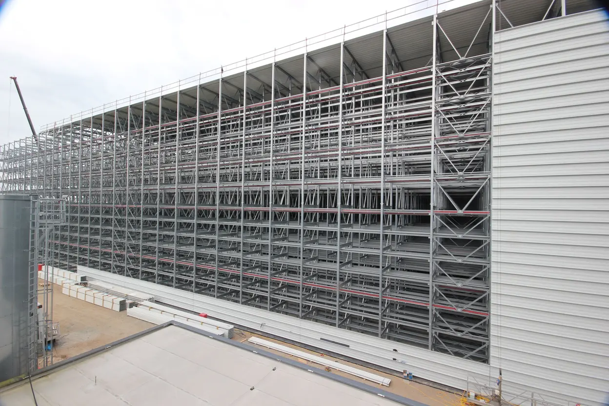 Ein großer, moderner Lagerbau mit einem beeindruckenden Stahlregalsystem, das die gesamte Fassade einnimmt. Der Platz ist teilweise leer, und einige Materialien liegen am Boden. Der Himmel ist bewölkt, was auf eine düstere Atmosphäre hinweist.