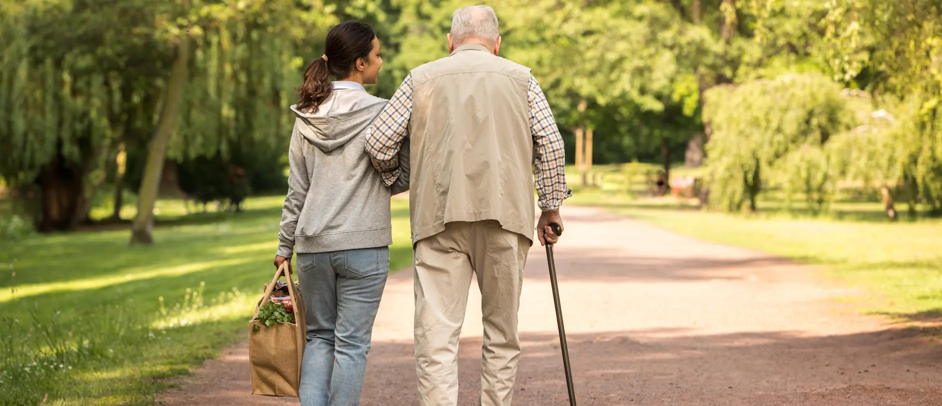 Eine junge Frau geht arm in arm mit einem älteren Mann auf einem schmalen Weg in einem Park. Der Mann stützt sich auf einen Gehstock, während die Frau eine Tasche mit Blumen trägt. Das Bild vermittelt eine Atmosphäre von Fürsorge und Verbundenheit.