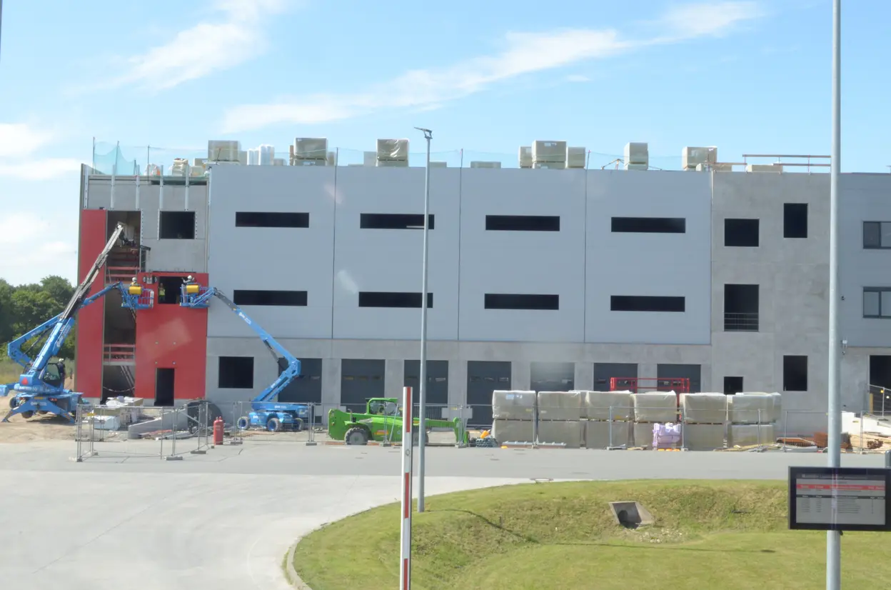 Eine Baustelle mit einem großen, dreigeschossigen Gebäude in grauer und roter Farbe. Bauarbeiter nutzen eine Scherenbühne, um an der Fassade zu arbeiten. Material wie Ziegel und Baustellenwagen sind in der Nähe sichtbar. Im Hintergrund sind Bäume und der Himmel zu sehen.