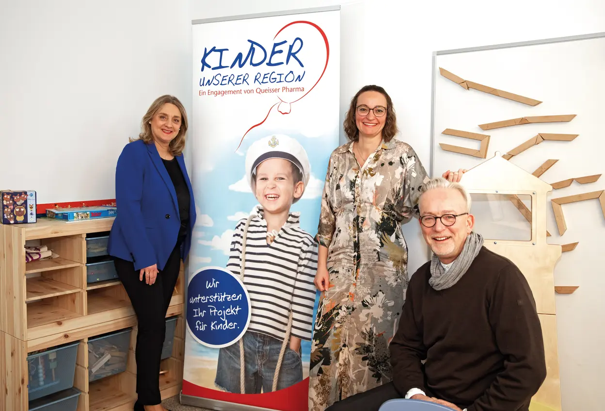 Mitarbeiter*innen von Queisser Pharma vor einem Roll-Up, das ein Kind in Matrosenverkleidung mit dem Logo "Kinder unserer Region" abbildet
