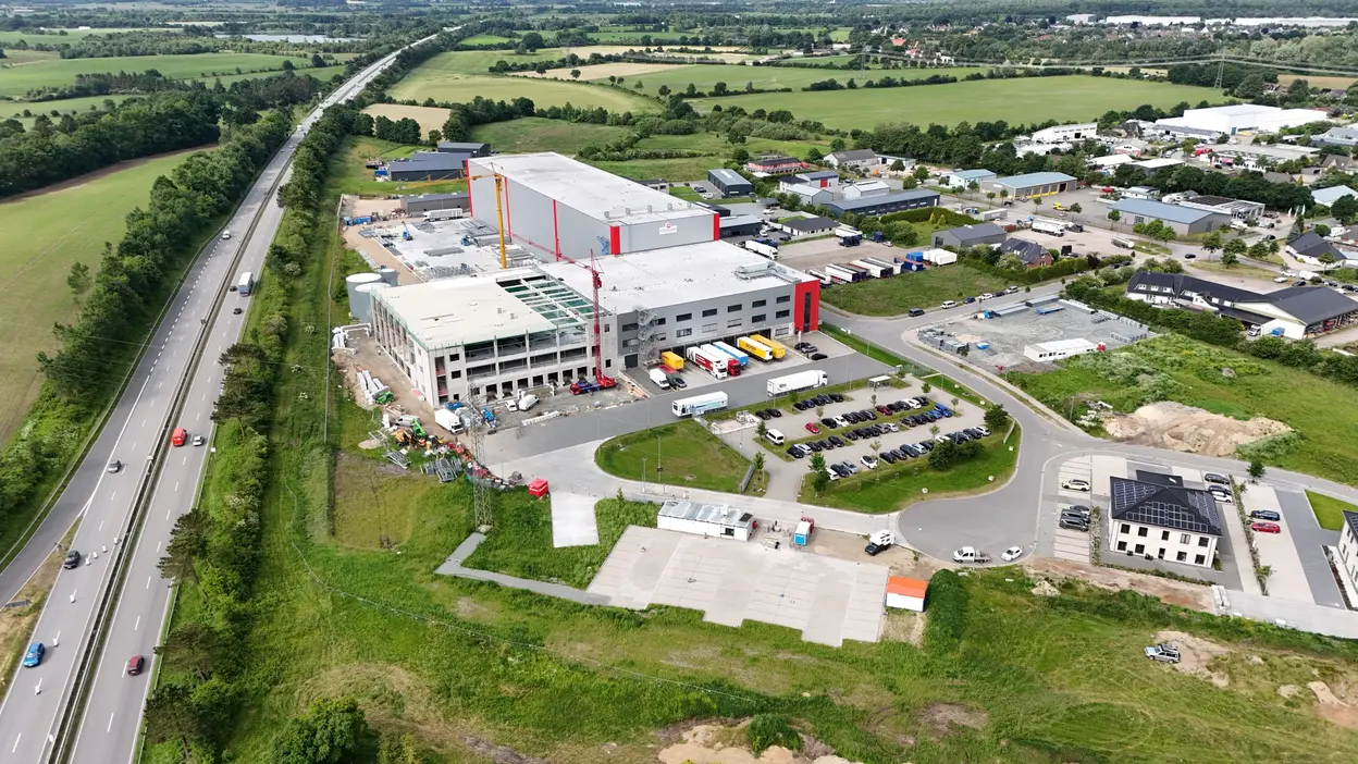 Eine Luftaufnahme zeigt ein großes, modernes Gebäude mit mehreren Lkw vor dem Eingang. Umgeben von einer grünen Landschaft und einer Straße, sind auch Parkplätze und weitere Gebäude in der Nähe sichtbar. Die Szene vermittelt den Eindruck eines industriellen Areals.