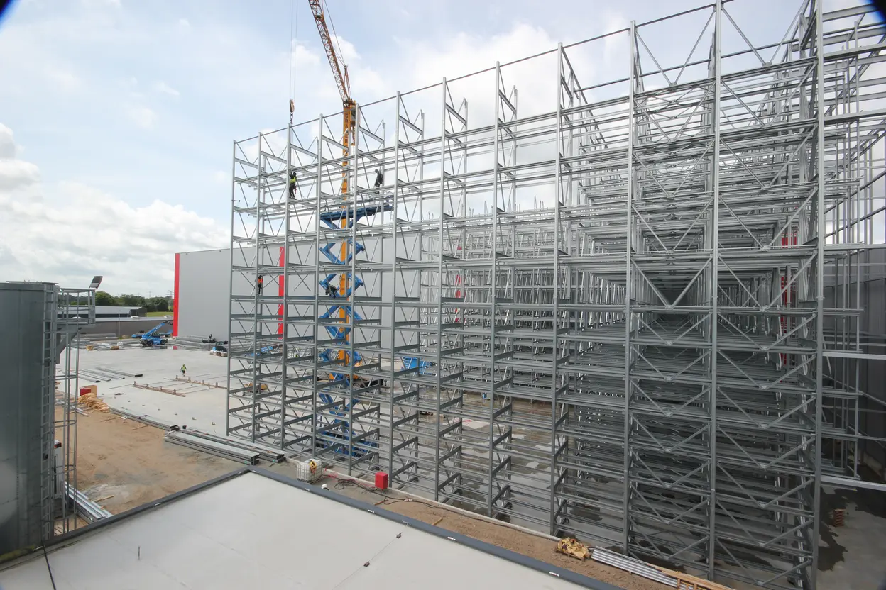 Der Bildinhalt zeigt eine Baustelle mit einem hohen, noch unvollständigen Stahlgerüst. Im Vordergrund ist ein Kran zu sehen, während Arbeiter an verschiedenen Stellen des Gerüsts aktiv sind. Der Boden ist teilweise bearbeitet und zeigt Materialien für den Bau.
