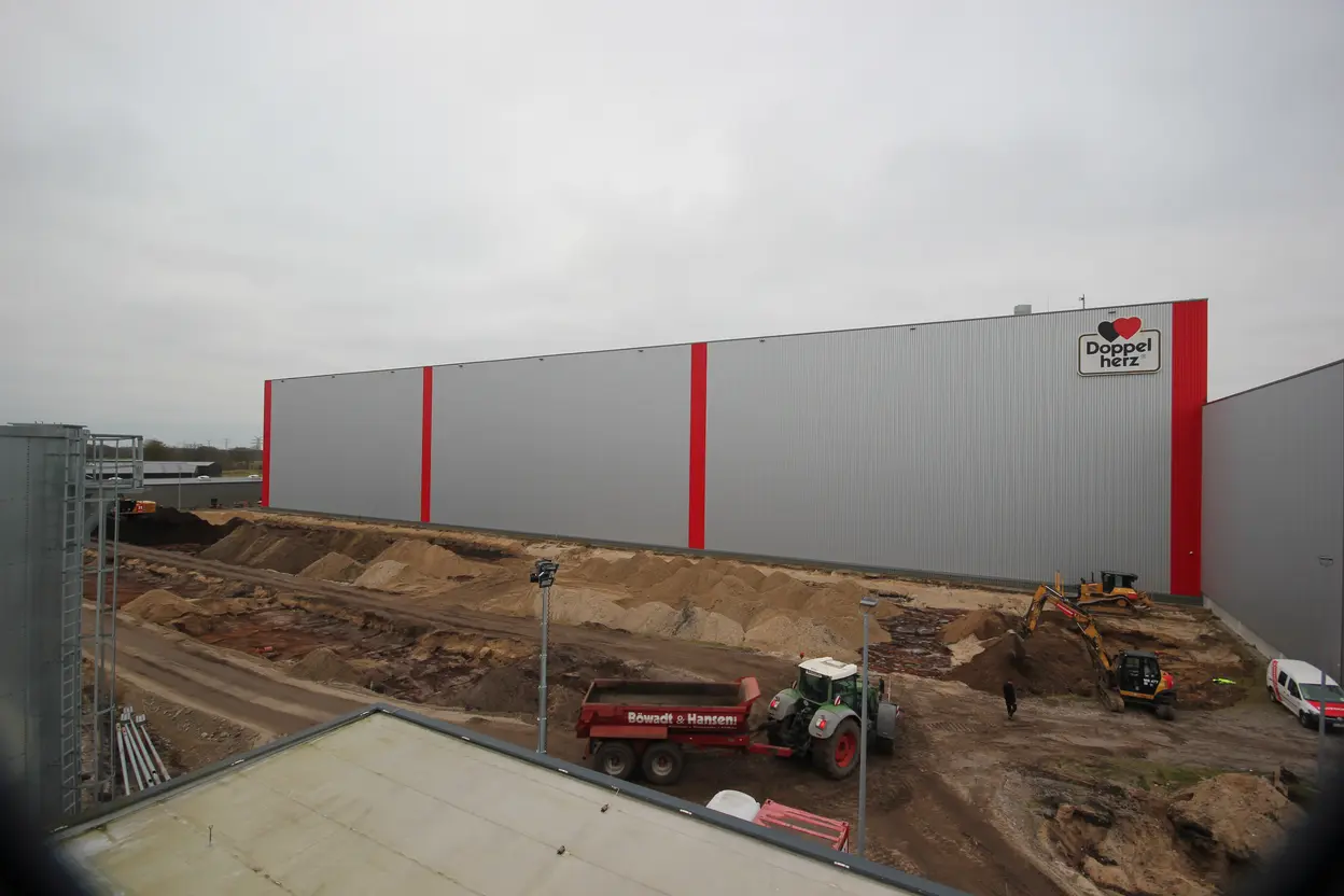 Baufortschritt am Queisser-Logistikzentrum mit Sandaufschüttung, Baggerarbeiten und LKW vor der Hallenfassade mit Doppelherz-Logo.
