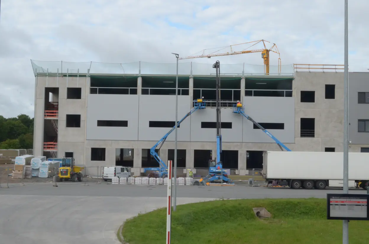 Auf dem Bild ist ein im Bau befindliches Gebäude zu sehen. Zwei Bauarbeiter auf einer Hebebühne arbeiten an der Fassadenverkleidung, während ein Kran in der Nähe steht. Im Vordergrund sind Baumaterialien und ein gelbes Baufahrzeug zu erkennen.