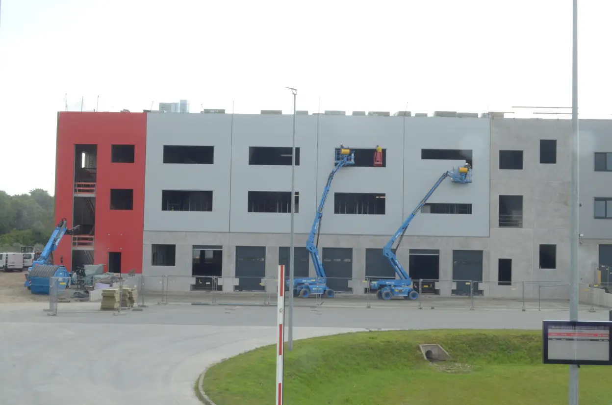 Ein Baustellenbild zeigt einen Gebäudekomplex im Bau. Arbeiter auf zwei hohen Hebebühnen führen Fassadenarbeiten durch. Die linke Gebäudeseite ist rot, die rechte grau. Unter den Arbeitsbühnen befinden sich Baumaterialien und Absperrungen.
