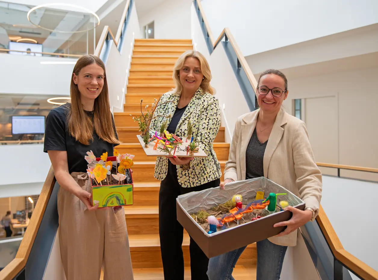 Drei Frauen stehen auf einer Treppe in einem modernen Gebäude und halten kreative Bastelarbeiten in ihren Händen. Sie lächeln und wirken freundlich. Im Hintergrund ist das Architekturdesign des Raumes sichtbar, mit einer offenen Treppe und einer hellen, einladenden Atmosphäre.