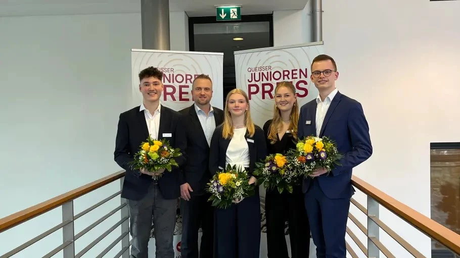 Fünf Personen (Preisträger*innen und Geschäftsführer Axel Kaempfe) stehen auf einem Galerieflur vor zwei Roll-Ups mit der Aufschrift ‚Queisser Junioren Preis‘; die vier Preisträger*innen halten Blumensträuße.