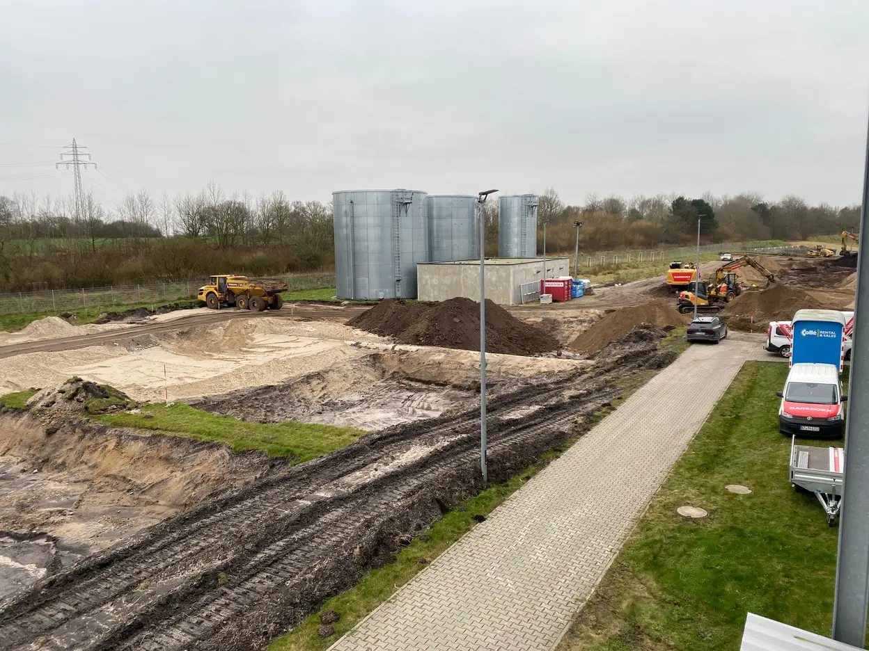 Baustellenszene mit Erdarbeiten, mehreren Baustellenfahrzeugen und drei großen Silos mit angrenzender Grünfläche.