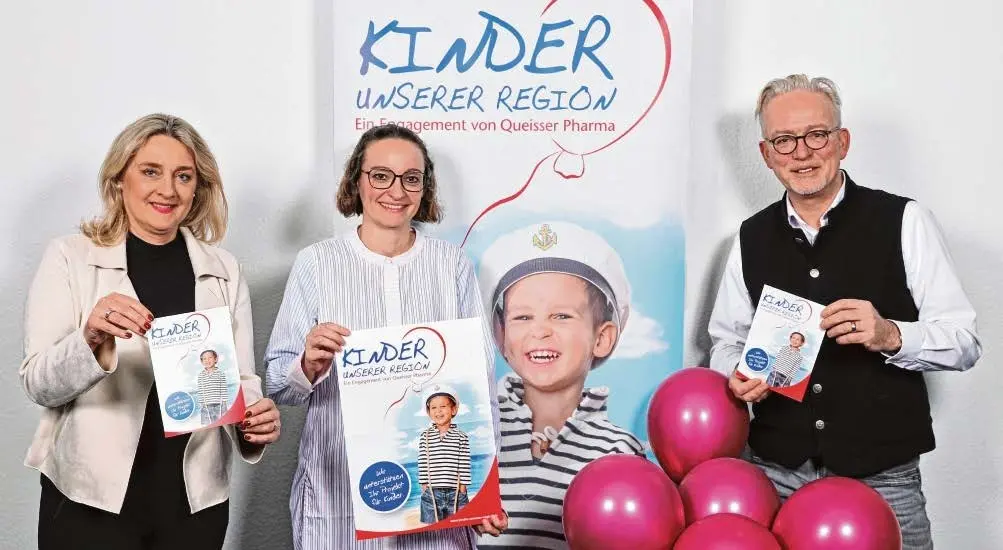 Drei Mitarbeiter*innen von Queisser Pharma stehen vor einem Roll-up mit der Aufschrift "Kinder unserer Region" und halten das gleichnamige Magazin in den Händen; im Vordergrund pinkfarbene Luftballons