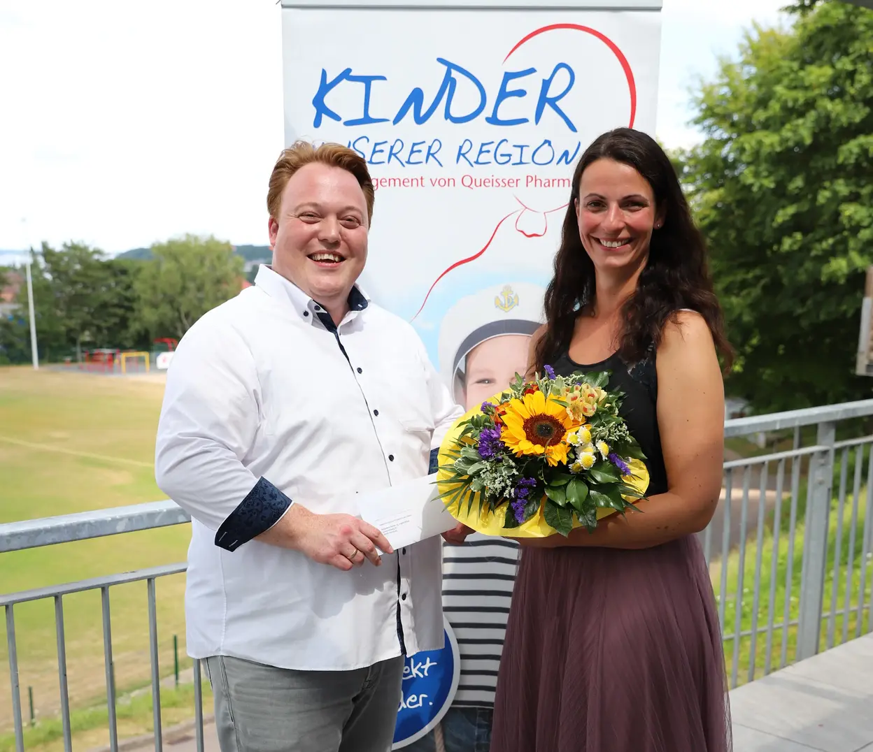 Zwei Personen stehen vor einem Roll-up mit der Aufschrift ‚Kinder unserer Region‘; eine Frau hält einen Blumenstrauß, der Mann hält einen Umschlag.“