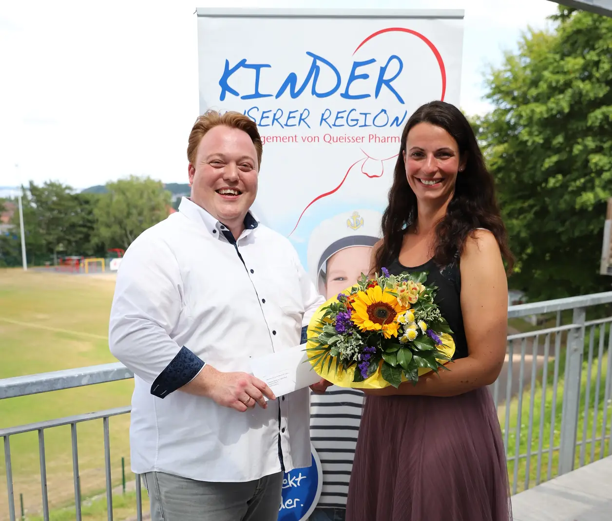 Zwei Personen stehen vor einem Roll-up mit der Aufschrift ‚Kinder unserer Region‘; eine Frau hält einen Blumenstrauß, der Mann hält einen Umschlag.“
