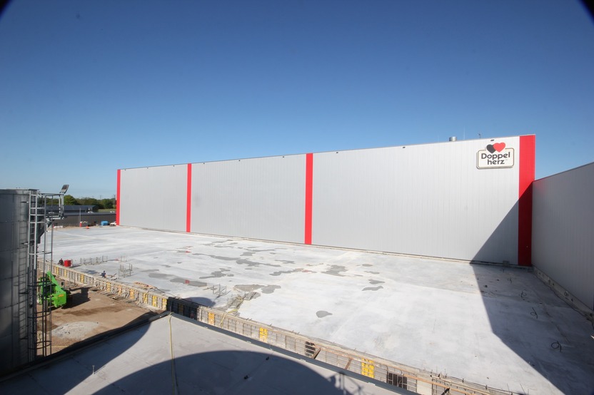 A large, gray warehouse building with red stripes stands against a clear blue sky. The foreground shows a construction area with concrete and equipment, indicating ongoing work at the site. The sign on the building displays the name "Doppel herz."