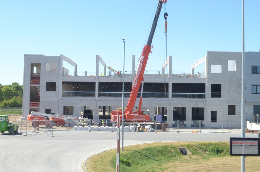 A construction site featuring a partially built building with concrete walls and open spaces. A large red crane is lifting materials, while construction equipment and piles of bricks are visible nearby. Workers in safety vests can be seen on the upper floors.