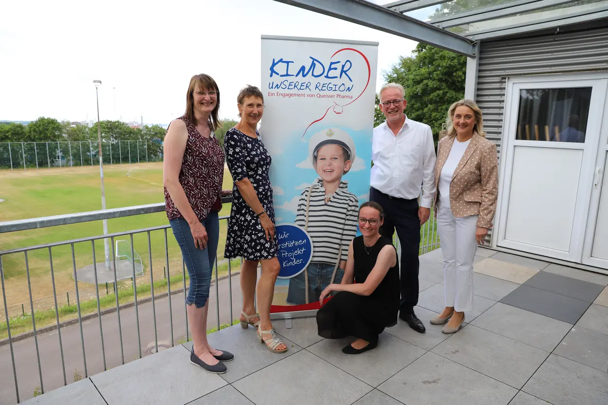 Vier Personen stehen und eine Person kniet neben einem Roll-up mit der Aufschrift "Kinder unserer Region" auf einer überdachten Terrasse mit Blick auf ein Sportfeld.
