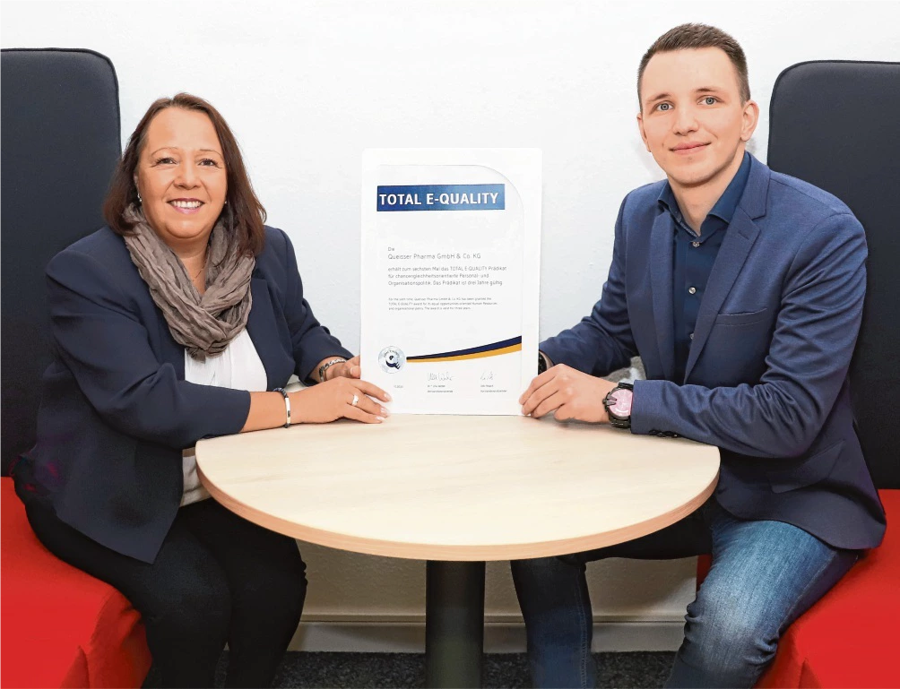 Zwei Personen sitzen an einem runden Tisch und halten gemeinsam eine Urkunde mit der Aufschrift ‚Total E-Quality‘.