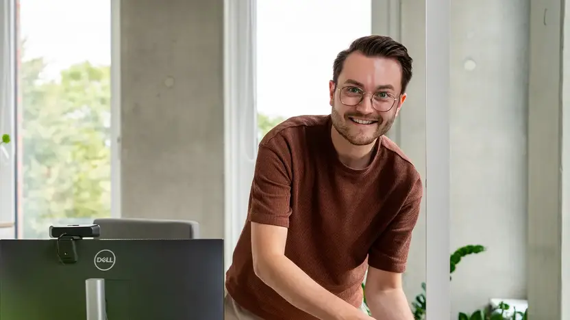 Junger Mann mit Brille lächelt im modernen Büro an seinem Arbeitsplatz.