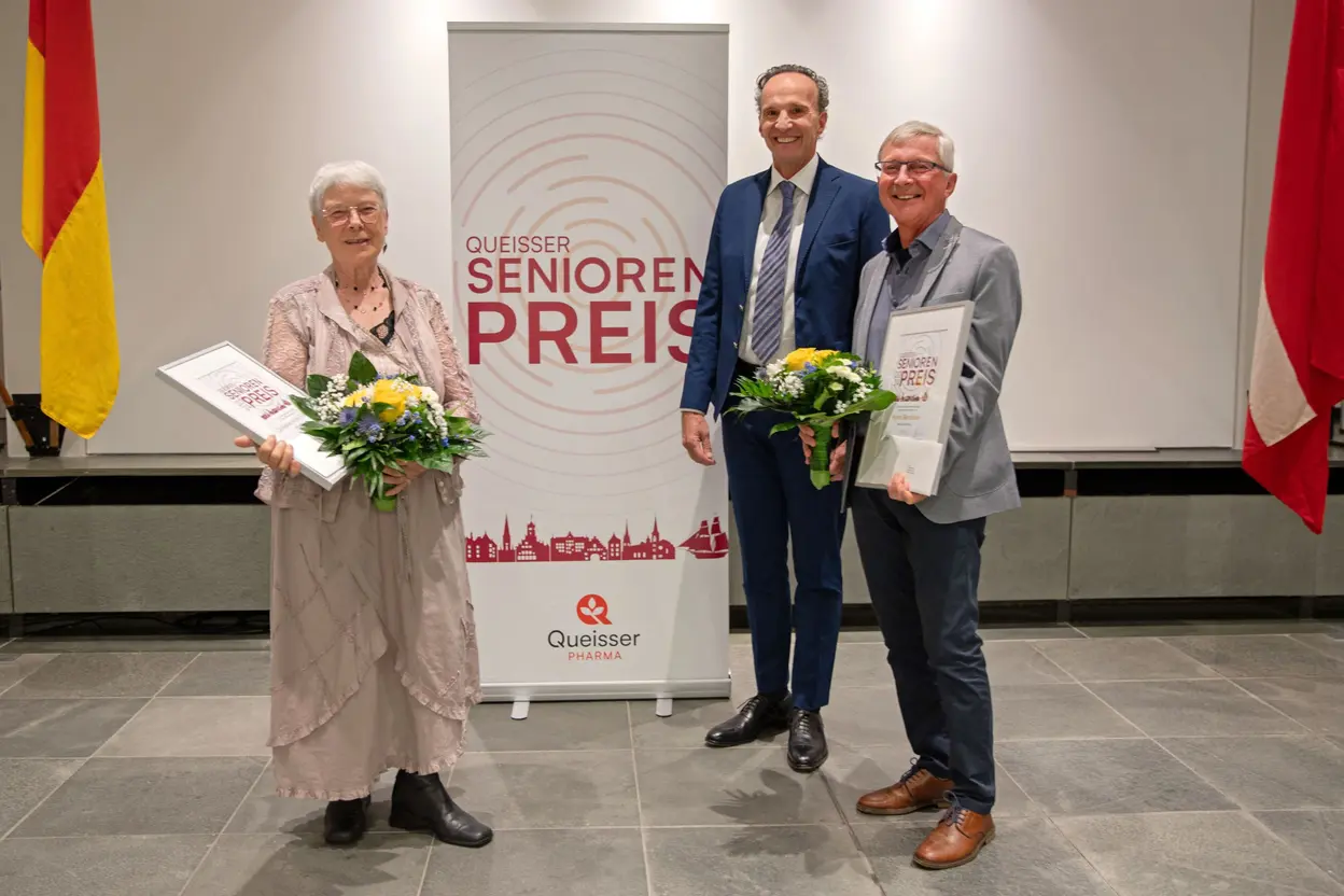 Eine Dame und zwei Herren vor einem Banner in feierlicher Umgebung