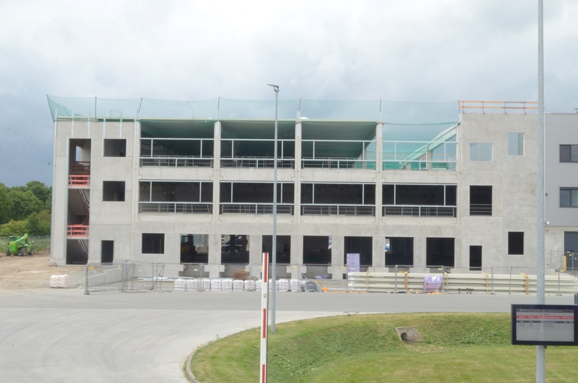 A partially constructed building is visible, featuring concrete walls and large openings for windows. The structure has several levels with scaffolding on one side. In the foreground, there are stacks of building materials and construction equipment, while the sky is overcast.