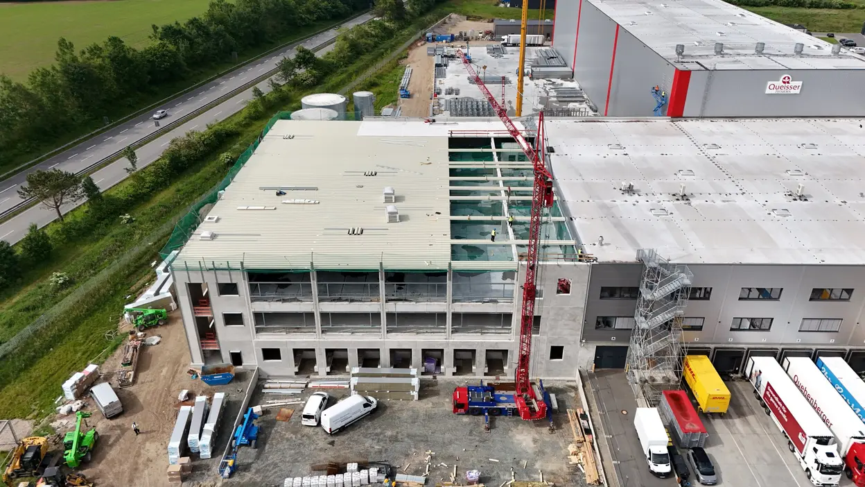 Ein Baustellenblick auf ein neues Gebäude mit einem roten Kran, der Materialien anhebt. Das Gebäude hat teilweise aufgerissene Wände und ist noch im Bau. Im Vordergrund sind Baustellenfahrzeuge und -materialien sichtbar, während sich eine Straße mit Fahrzeugen an der Seite des Geländes erstreckt.