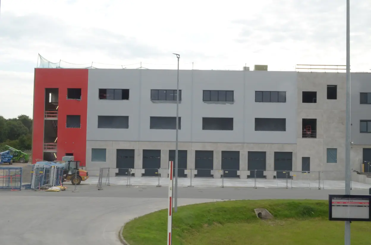Die Abbildung zeigt ein im Bau befindliches Gebäude mit einer roten und grauen Fassade. Einige Fenster sind bereits eingesetzt, während andere noch offene Baustellen sind. Im Vordergrund sind Baustellenelemente und eine leicht geschwungene Straße zu sehen.