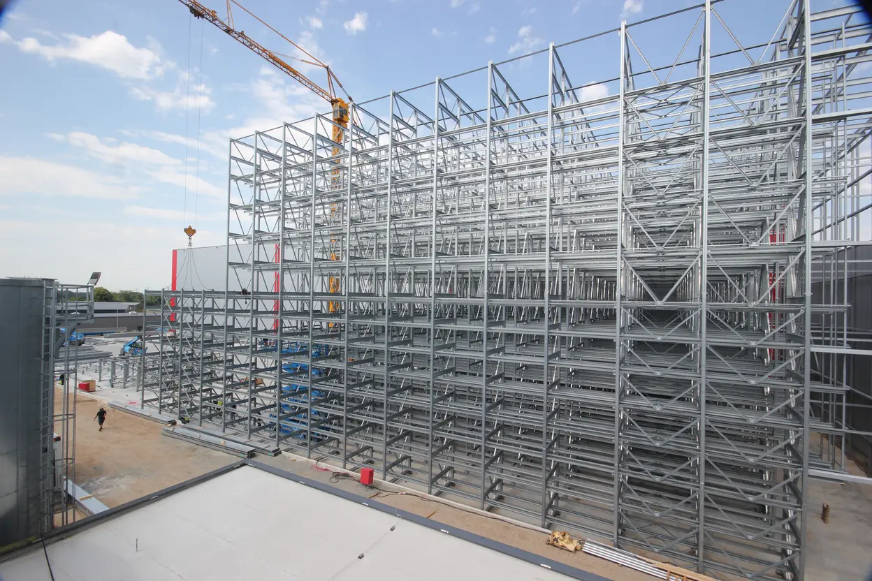 Das Bild zeigt ein großes Gerüst aus Stahlrahmen auf einer Baustelle, teilweise im Bau. Im Hintergrund ist ein Baukran zu sehen, der Materialien hebt. Ein Arbeiter bewegt sich im Vordergrund. Der Himmel ist klar mit wenigen Wolken.