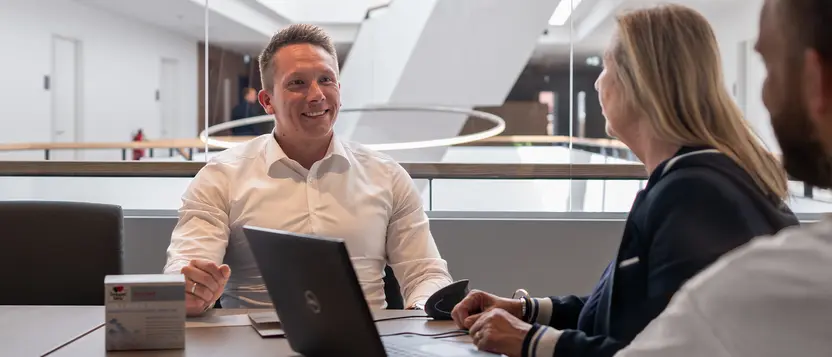 Drei Personen im Gespräch an einem Besprechungstisch mit Laptop und Produktverpackung.