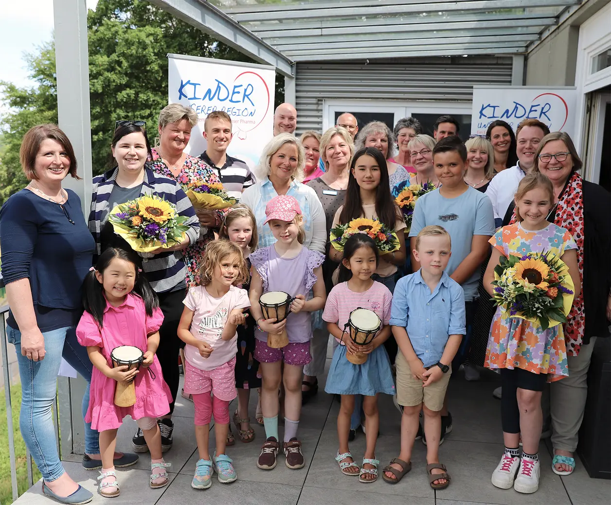 Gruppenfoto mit Kindern und Erwachsenen auf einer überdachten Terrasse vor Roll-ups mit der Aufschrift ‚Kinder unserer Region‘; Kinder halten Trommeln, Erwachsene Blumensträuße.
