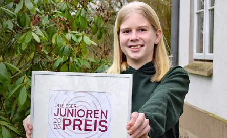 Ein Mädchen mit langen, blonden Haaren hält stolz ein Schild mit der Aufschrift „QUER ISSER JUNIOREN PREIS 2025“. Der Hintergrund zeigt eine Wand und grüne Pflanzen.
