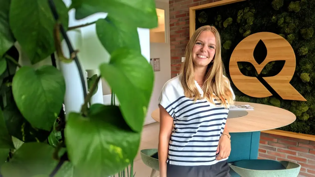 Eine lächelnde Frau mit langen, blonden Haaren trägt ein gestreiftes Poloshirt. Sie steht neben einem Tisch in einem modernen Raum, an der Wand ist ein großes, pflanzenbasiertes Logo zu sehen. Grüne Pflanzen sind im Vordergrund sichtbar.