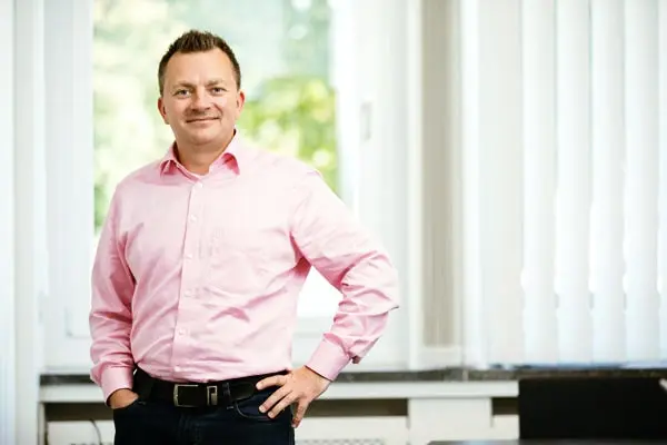 Freundlich lächelnder Mann in einem rosa Hemd steht vor einem Fenster