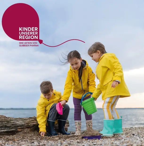 Drei Kinder in gelben Regenjacken spielen am Strand. Ein Junge sammelt Sand mit einer Schaufel, während zwei Mädchen mit Eimer und Schaufel ebenfalls im Sand graben. Die Szenerie zeigt einen bewölkten Himmel und das Wasser im Hintergrund.