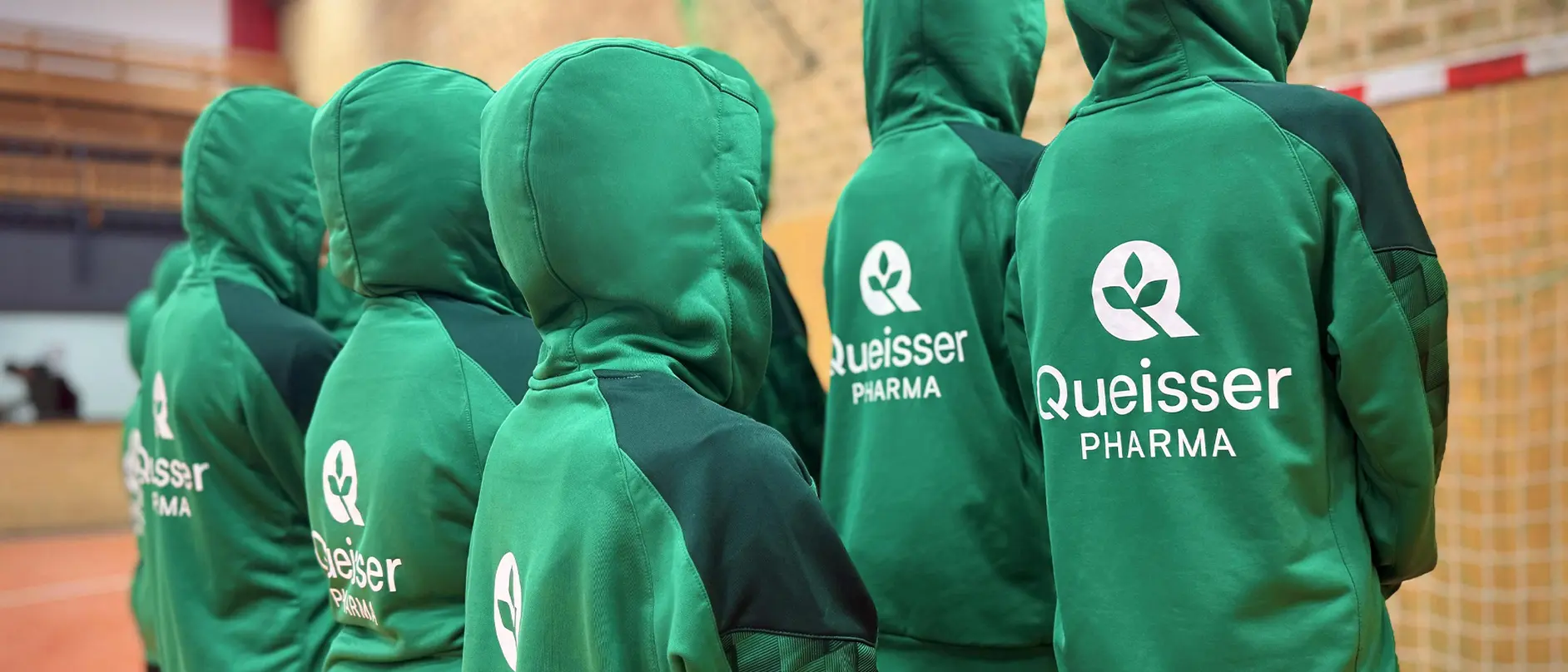 Sportteam mit grünen Kapuzenpullovern, auf dem Rücken das Queisser-Logo, von hinten fotografiert in einer Sporthalle.