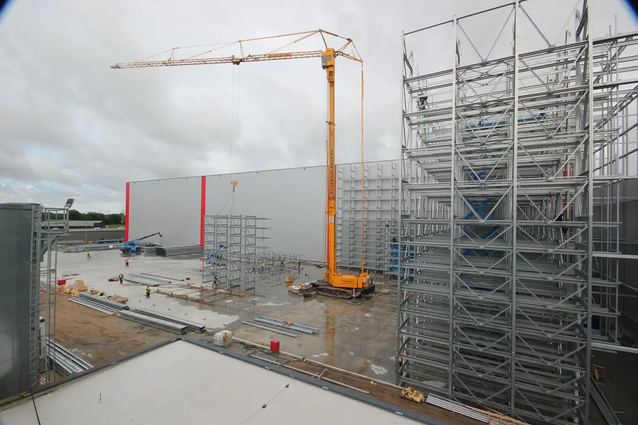 Eine Baustelle mit einem großen Kran, der ein Gerüst hebt. Im Hintergrund sind weitere Bauarbeiten sichtbar, darunter Mitarbeiter in Sicherheitskleidung und Materialien auf dem Boden. Der Himmel ist bewölkt, und es sind zwei große, graue Gebäude zu erkennen.