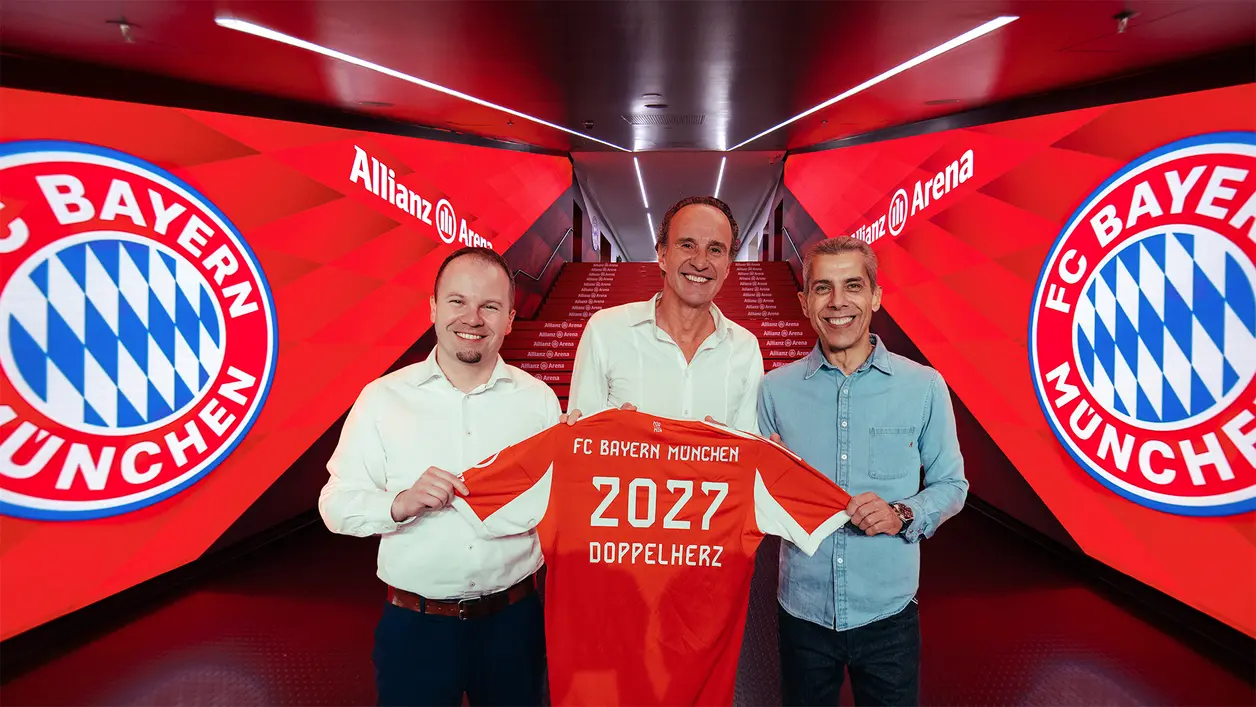 Drei Männer stehen in der Allianz Arena und halten ein rotes Trikot des FC Bayern München mit der Aufschrift "2027 Doppelherz". Im Hintergrund sind die Logos des Vereins und eine symmetrische, rot gestaltete Wand sichtbar.