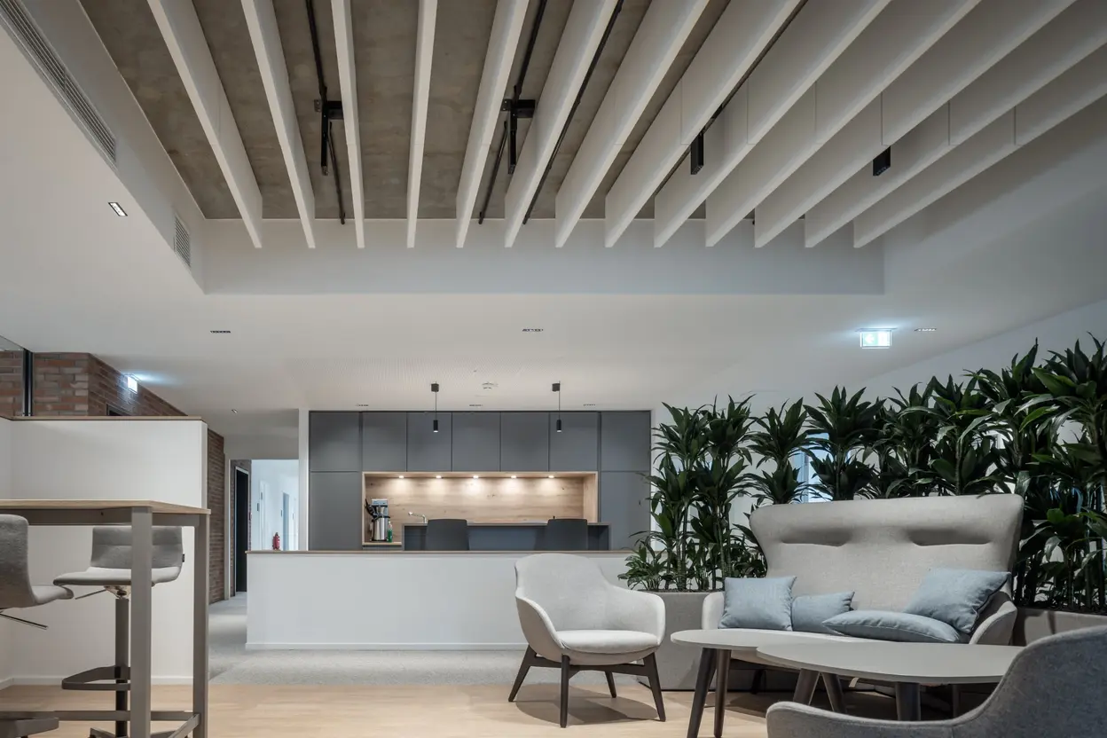 Modern gestalteter Lounge-Bereich mit Sesseln, Couch und Couchtisch vor einer begrünten Raumtrennung, im Hintergrund offene Teeküche und Holzdecke