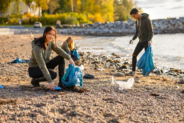 Drei Menschen sammeln Müll am Strand. Eine Frau sitzt am Boden und hebt Abfälle in eine blaue Tüte, während zwei weitere Personen im Hintergrund den Strand reinigen. Die Umgebung zeigt naturbelassene Elemente in einer herbstlichen Szenerie.