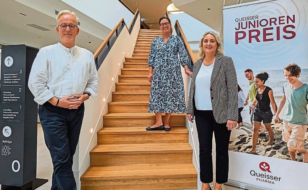 Drei Personen stehen an einer Holztreppe im Queisser-Gebäude neben einem Roll-up mit der Aufschrift ‚Queisser Junioren Preis‘ und Jugendlichen am Strand.