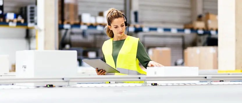 Mitarbeiterin im Lager mit Warnweste prüft Ware und hält ein Tablet.