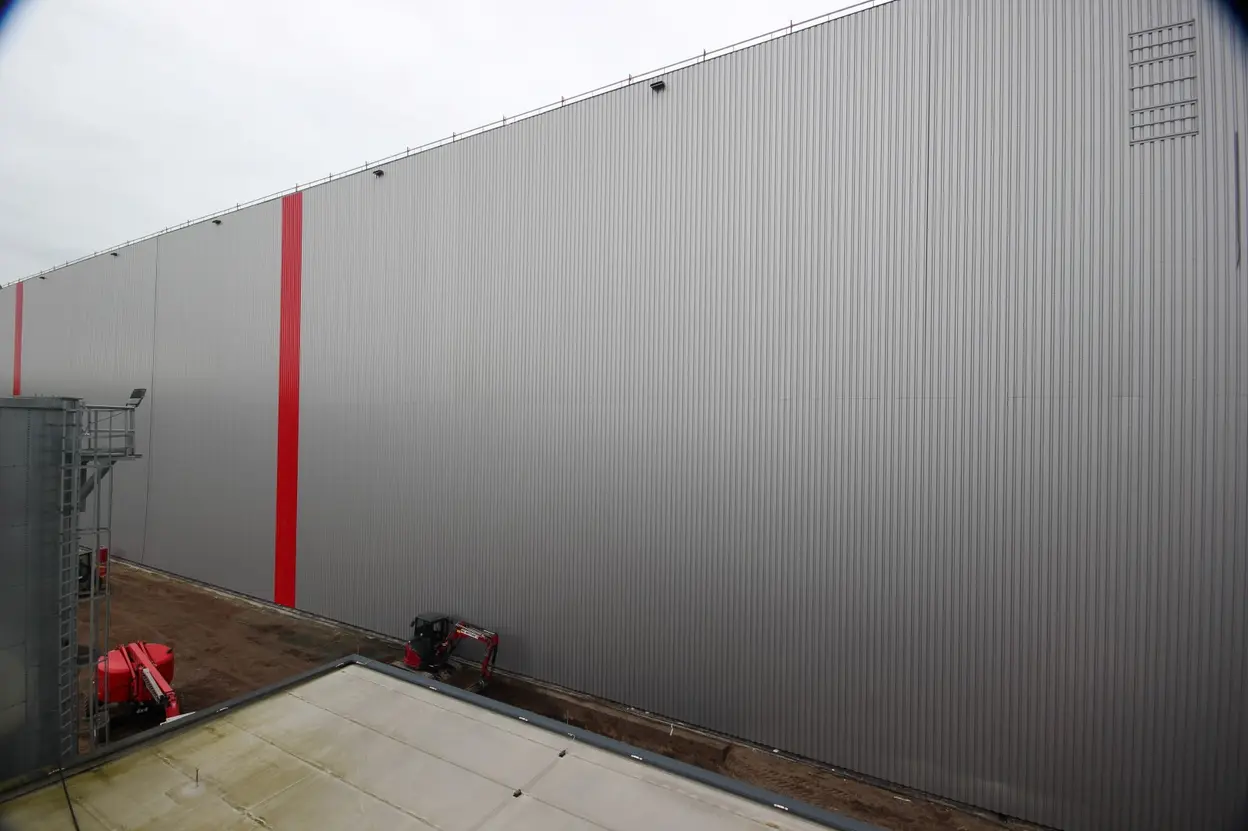 Eine große, graue Lagerwand mit einer vertikalen roten Streife. Der Himmel ist bewölkt. Im Vordergrund sind Teile einer anderen Struktur zu sehen, die den Boden an der Basis der Wand berührt. Die Umgebung wirkt industriell und strkt.