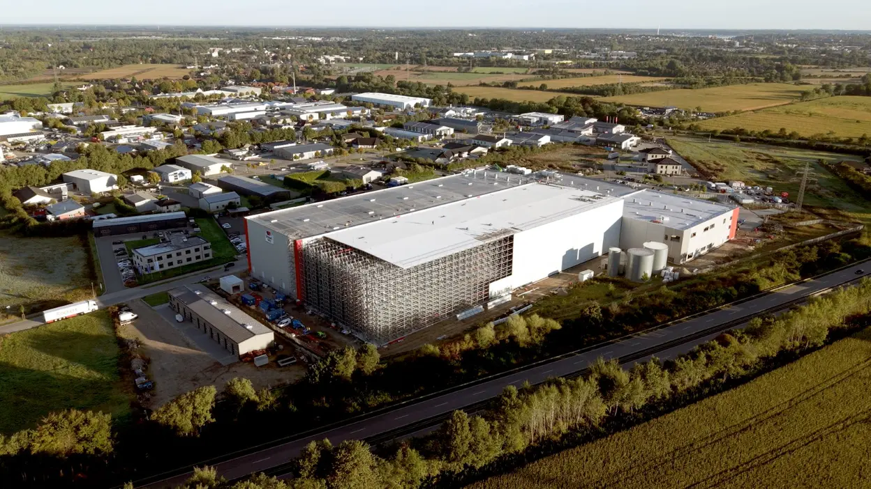 Die Aufnahme zeigt ein großes Lagergebäude in einem industriellen Gebiet, umgeben von weiteren Gebäuden und landwirtschaftlichen Flächen. Der Fokus liegt auf dem modernen, weitläufigen Lager mit einem flachen Dach und einer teilweise sichtbaren Außeneinrichtung. Im Hintergrund sind Bäume und weitere Strukturen zu sehen.