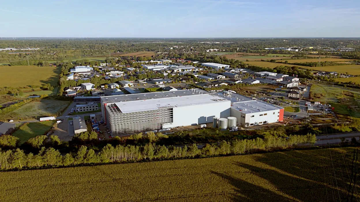 Luftaufnahme eines großen Gebäudes, umgeben von Feldern und einer kleinen Stadt. Die Struktur zeigt verschiedene Anbauten und Fenster. Im Vordergrund sind grüne Felder zu sehen, hinter dem Gebäude befinden sich Wohn- und Industriegebäude. Heller Himmel mit wenigen Wolken.