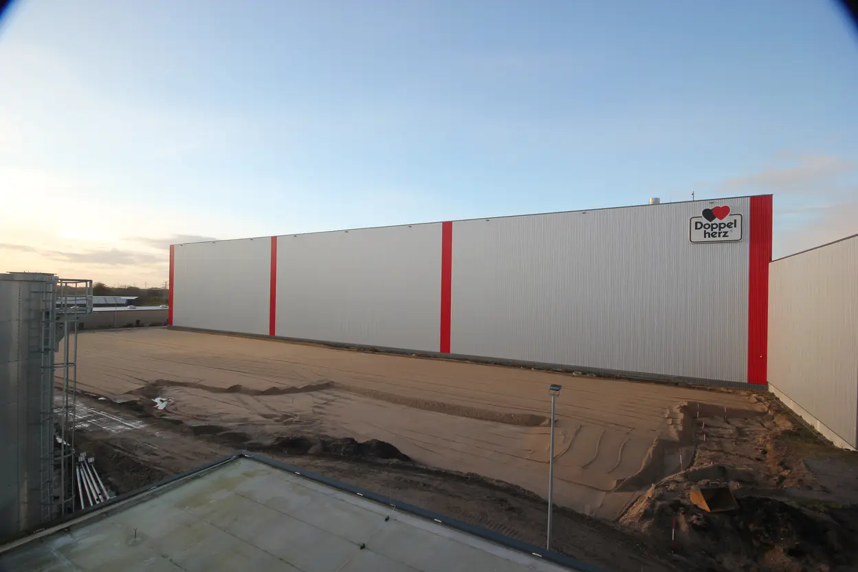 Vorbereitete Baufläche vor der Fassade des Queisser-Logistikzentrums mit rotem Doppelherz-Logo, planiertes Sandbett für nachfolgende Baumaßnahmen