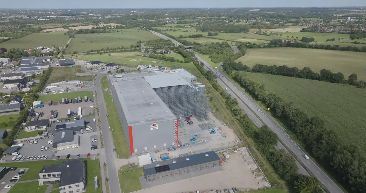 Eine Luftaufnahme eines großen, modernen Industriegebäudes mit einem grauen Dach und roter Seite. Im Vordergrund sind mehrere Parkplätze und Fahrzeuge zu sehen. Die Umgebung ist von grünen Feldern und Straßen umgeben, die in der Nähe des Bauwerks verlaufen.