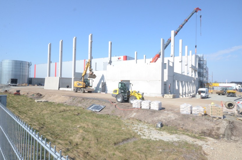 Construction site with heavy machinery, including a crane and excavators, actively working on a building. Visible are concrete foundations and materials stacked on the ground, with a backdrop of a partially completed structure under a clear blue sky.