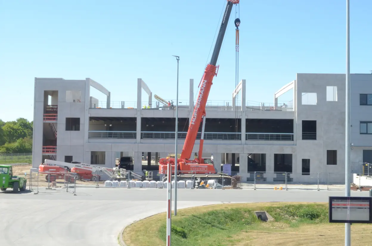 A construction site featuring a partially built building with concrete walls and open spaces. A large red crane is lifting materials, while construction equipment and piles of bricks are visible nearby. Workers in safety vests can be seen on the upper floors.